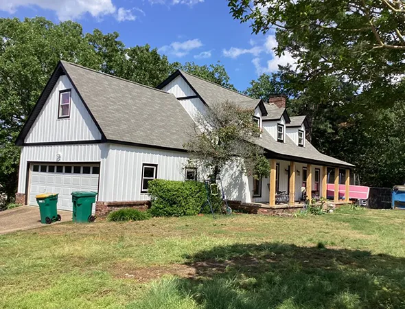 A two-story house with gray shingle roofing and white siding stands amid green trees. The front porch has columns, and two green bins sit near the garage.