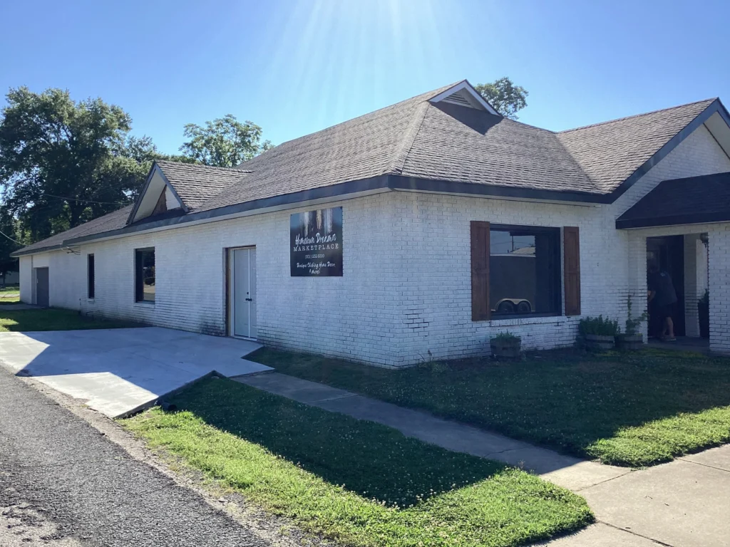 White brick building with a gabled roof on a sunny day. A sign reads "Heather Renee’s Marketplace." Large windows and a small yard add charm.