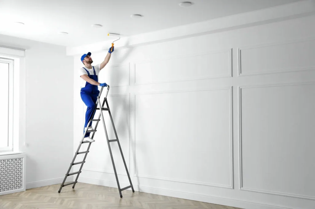 A person in blue overalls and cap paints a white ceiling while standing on a ladder in a bright, empty room, conveying a sense of focus and precision.