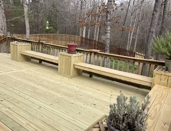 A spacious wooden deck features built-in benches and planters. A red pot sits atop a bench. Bare trees and a forested backdrop suggest winter.