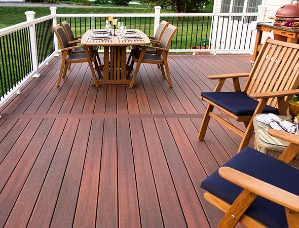 A wooden deck features a dining table set for four, with neatly arranged plates and glasses. Cozy chairs with blue cushions add a welcoming, elegant touch.