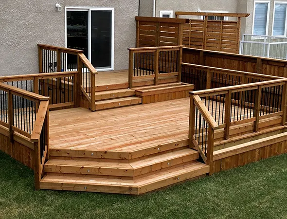Wooden deck with black railings, steps leading up, and a privacy screen against a house. The deck sits on a grassy lawn, creating a warm, inviting outdoor space.