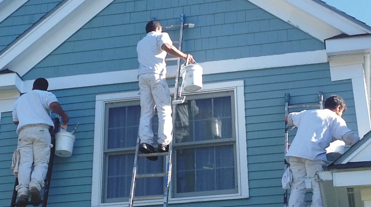 Three painters in white uniforms are on ladders painting the exterior of a blue house. The scene is bright and suggests teamwork.