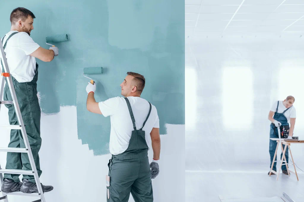 Two painters in overalls use rollers to apply teal paint on a wall. One stands on a ladder while the other works beside him. A third person in the background uses tools on a table in a bright, minimalist room.