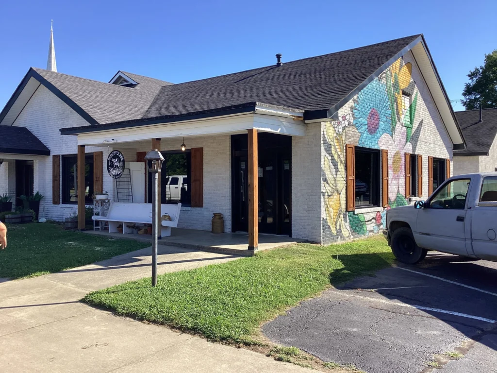 A white brick building with a colorful flower mural on the wall. A porch with wood accents and a bench sits under a bright blue sky. A pickup truck is parked nearby.