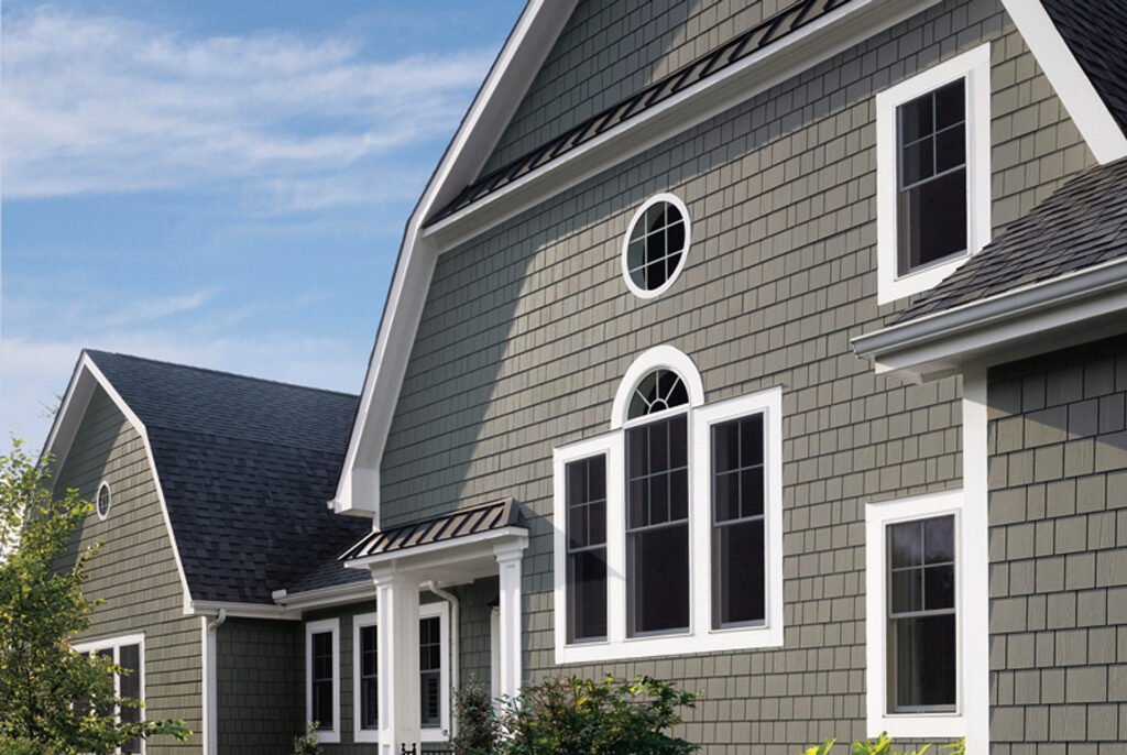 Modern gray house with white trim, featuring gabled roofs, large arched and circular windows. Sunny day, clear sky, lush greenery in foreground.