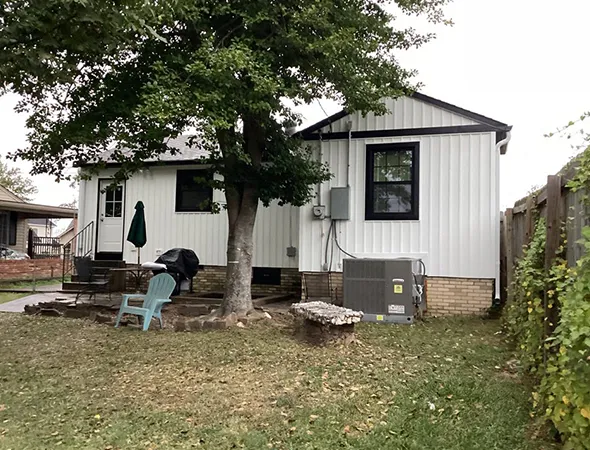 Small white house with black trim, surrounded by a grassy yard. A tree and outdoor seating, including blue chairs, add a cozy, peaceful atmosphere.