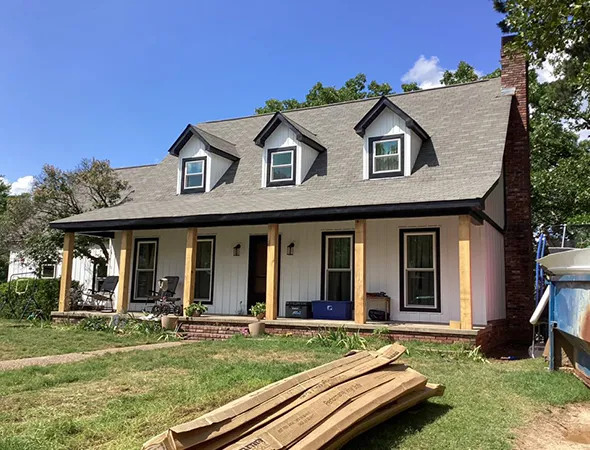 A white two-story house with a gray shingled roof and three dormer windows. A porch with wooden posts, and cardboard debris on a slightly overgrown lawn under a clear blue sky.