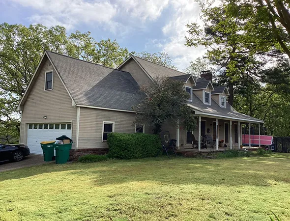 A beige two-story house with a sloped roof, dormer windows, and a wraparound porch. Surrounded by trees, the scene is calm and inviting