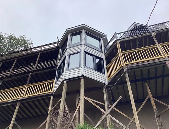 Elevated house with multiple wooden decks and a protruding bay window, supported by tall wooden stilts. The overcast sky adds a precarious tone.