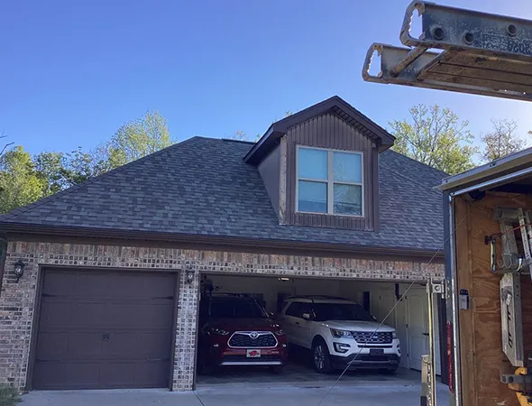 A two-story house with a brick facade and a prominent gabled roof is shown. The open garage reveals a red and a white car inside. A section of a ladder and a vehicle with tools is visible on the right, suggesting ongoing maintenance work. The scene is under a clear blue sky.