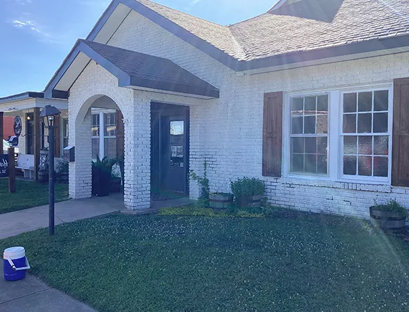 A bright, sunny view of a quaint white brick house with a green door, wooden shutters, and small plants by the entrance. A blue sky enhances the peaceful atmosphere.