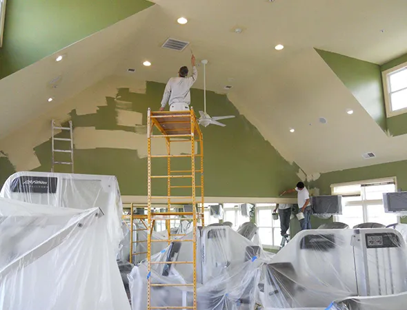 Two painters work in a gym, covering walls with light beige paint. Equipment is wrapped in protective plastic. The room feels calm and industrious.