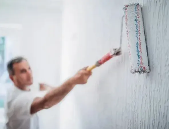 A person painting a white wall with a roller brush, focusing on smooth strokes. The setting is bright, conveying a sense of freshness and renewal.