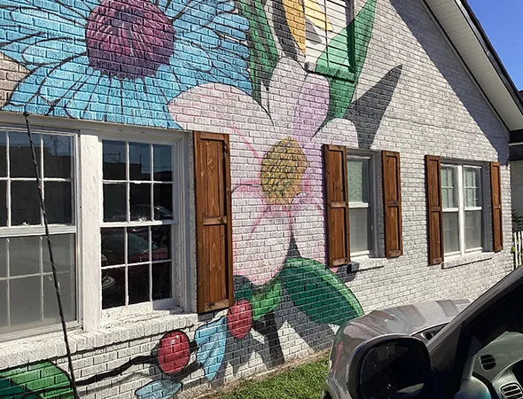 A white brick building with a colorful flower mural on the wall. A porch with wood accents and a bench sits under a bright blue sky.