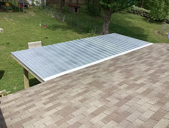 Rooftop view showing a metal patio cover with corrugated sheets next to a shingled roof. A grassy backyard with trees is visible in the background.
