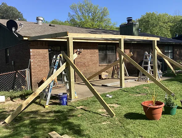 Brick house with partially constructed wooden pergola in the backyard. Ladders and tools are scattered around on a sunny day, with potted plants nearby.