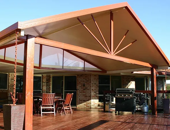 Spacious wooden patio with modern gabled roof, outdoor furniture, barbeque grill, and flower pot. Bright sunny day emphasizes cozy ambiance.