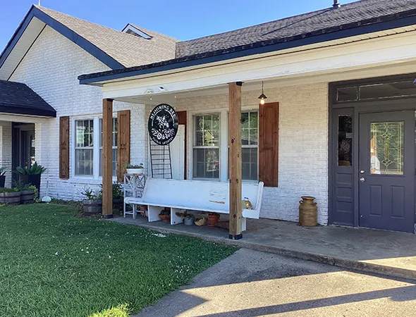White brick house with wooden shutters, a covered porch, and a dark gray door. A vintage sign and bench enhance the cozy, rustic charm.