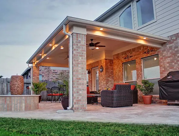 Covered patio with brick pillars and a ceiling fan, furnished with wicker chairs and a grill. Warm lighting creates a cozy evening ambiance.