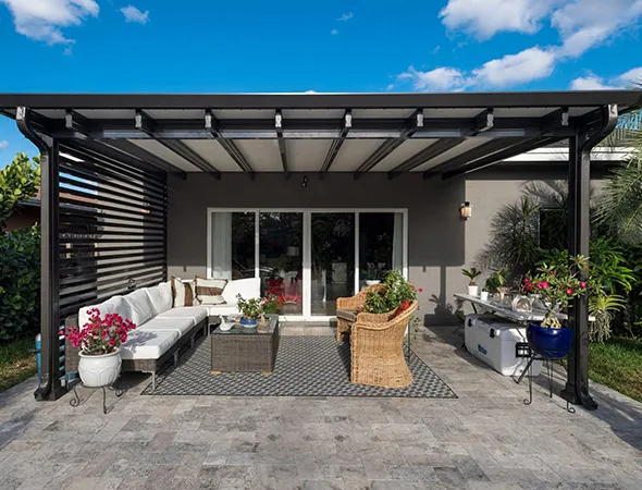 A modern patio with a pergola, featuring a white sectional sofa, wicker chairs, plants, and a rug on a stone floor. The scene is bright and inviting.