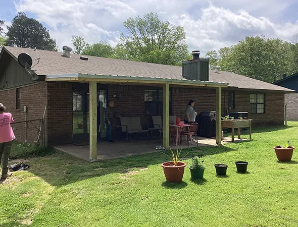 Backyard scene with a brick house, a covered patio, and people relaxing. Potted plants are on the grassy yard, and a cloudy sky is overhead. Casual, serene setting.