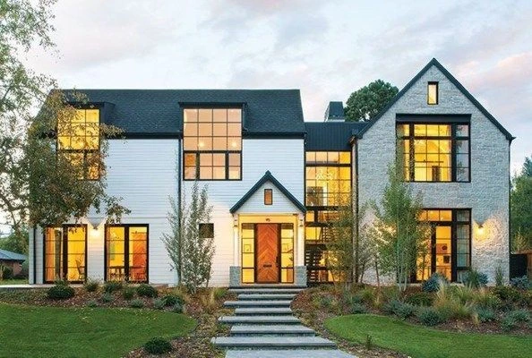 Modern farmhouse with a mix of white siding and stone exterior. Large, illuminated windows, lush landscaping, and a central path create a warm, inviting feel.