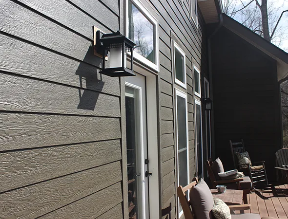 Modern house exterior with dark wood siding, featuring a row of tall windows and doors. A lantern casts a shadow, and chairs sit on a sunlit patio.