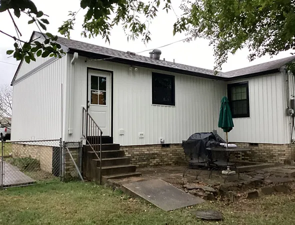 Small white house with a brick foundation, surrounded by a grassy yard. A door with steps leads to a patio with a table, chairs, and a green umbrella.