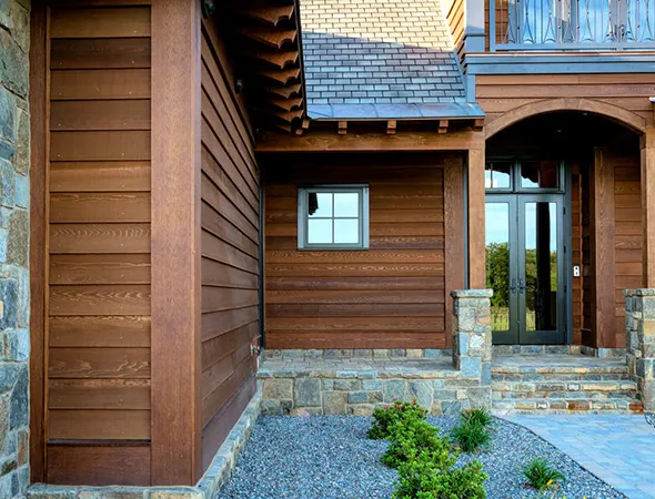 Modern house entrance featuring wood siding and stone accents. A glass door and small window add elegance. Gravel path with green plants in front. Rustic yet inviting.