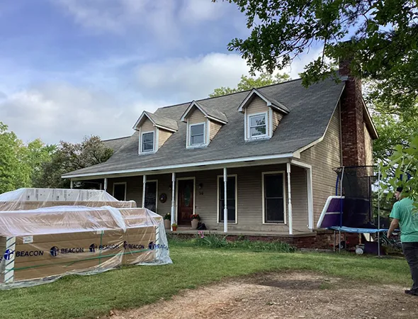 A two-story house with a gray sloped roof and dormer windows. Construction materials are stacked in the front yard, indicating ongoing renovations.