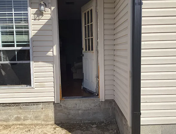 A partially open white door welcomes you into a home with beige siding, a large window to the left, and sunlight casting a warm glow across the exterior wall.