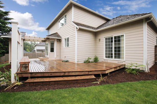 A beige two-story house with a wooden deck extends into a lush green lawn. The sky is partly cloudy, giving a fresh, serene ambiance to the setting.