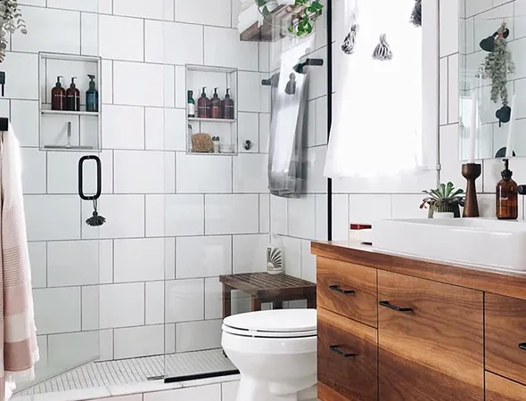 Bright bathroom with white tiles, wooden vanity, and potted plants. Glass shower with black fixtures. Natural light from window, cozy and modern.